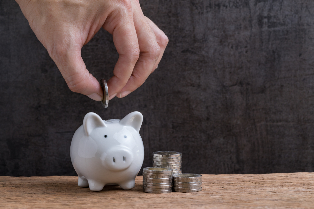 Man Hand Putting Coin Into Piggy Bank With Stack Of Coins close by