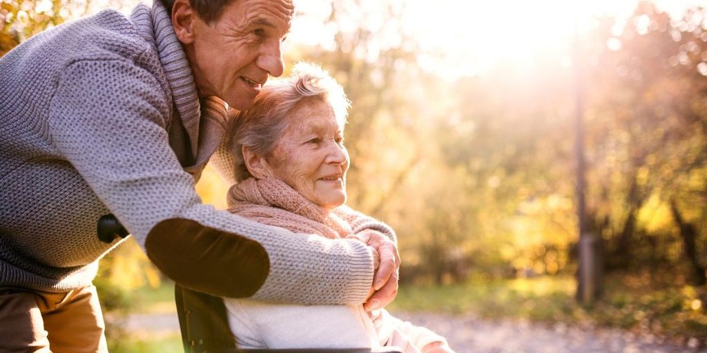 Senior,Man,And,Elderly,Woman,In,Wheelchair,In,Autumn,Nature. Man with elderly mother in wheelchair smiling behind her